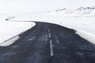 Пустая асфальтовая дорога уходит в заснеженную равнину, вдоль обочины жёлтые столбики
